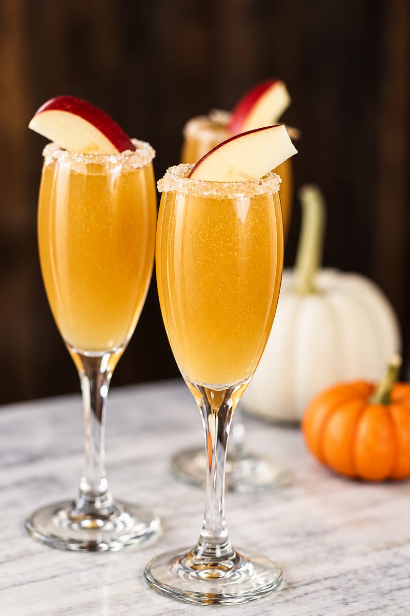 Three champagne flutes filled with a golden Apple Cider Mimosa cocktail, garnished with apple slices and sugar rims, sit on a marble surface with white and orange pumpkins in the background.