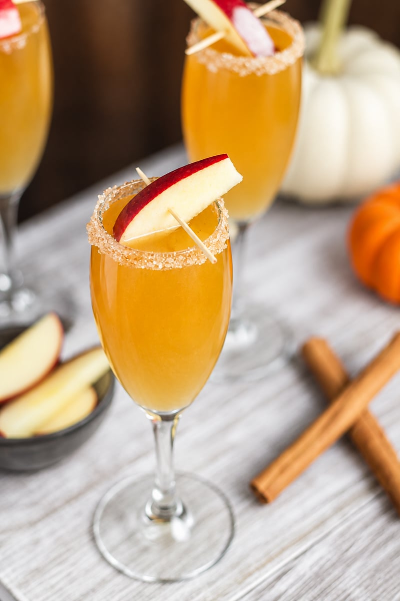 A champagne flute filled with an orange Apple Cider Mimosa, rimmed with brown sugar and garnished with apple slices. Cinnamon sticks and pumpkins are nearby on a wooden surface.