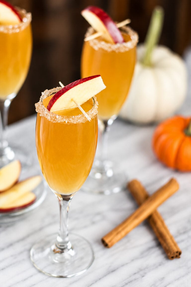Three glasses of autumn cocktails, including a festive Apple Cider Mimosa, are garnished with apple slices and brown sugar rims on a marble surface, with cinnamon sticks, apple slices, and small pumpkins in the background.