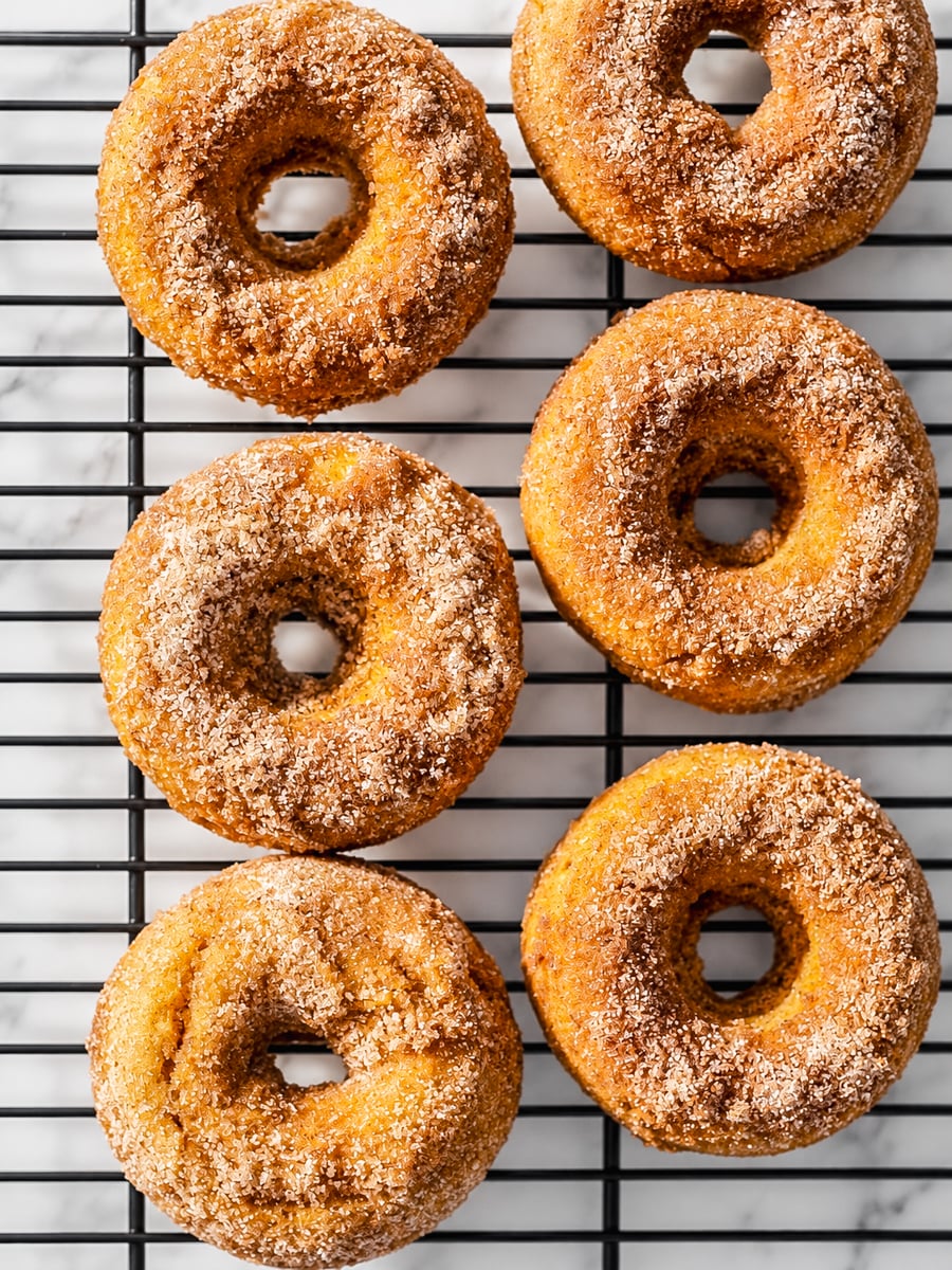 Six cinnamon sugar baked pumpkin spice donuts are arranged in two columns on a black wire cooling rack, placed over a white marble surface.