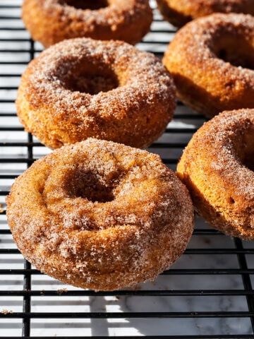 Six baked pumpkin spice donuts, coated in cinnamon sugar, are cooling on a black wire rack, with sunlight highlighting their texture and casting shadows on the marble surface below.