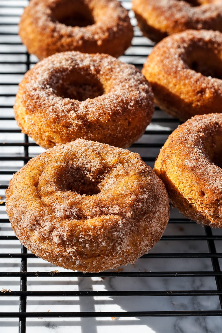 Six baked pumpkin spice donuts, coated in cinnamon sugar, are cooling on a black wire rack, with sunlight highlighting their texture and casting shadows on the marble surface below.