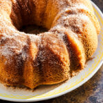 A golden-brown donut bundt cake dusted with cinnamon sugar on a white plate with a yellow rim.