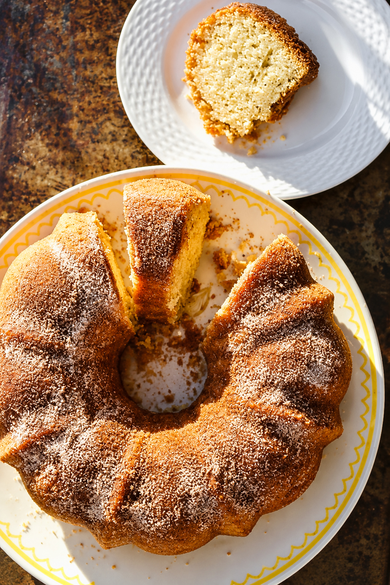 A sugar-dusted donut bundt cake on a patterned plate, with a single slice on a separate white plate.