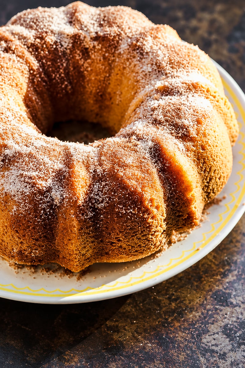 A golden-brown donut bundt cake dusted with cinnamon sugar on a white plate with a yellow rim.