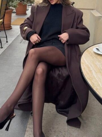 A person sits at an outdoor café table in Italy in December, wearing a long brown coat, black sweater, short skirt, sheer black tights, and black heels. A black handbag and a white cup rest on the table. The person's face is not visible.