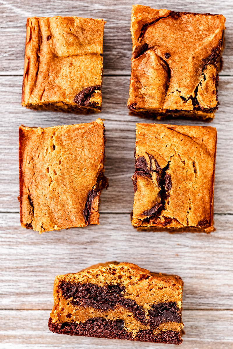 Five square pieces of pumpkin blondie on a wooden surface, with one piece showing its layered interior.