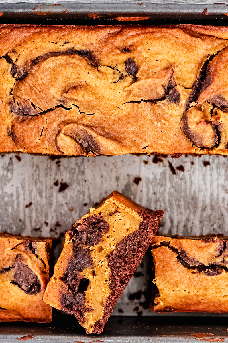A loaf of marbled chocolate pumpkin bread, similar in style to pumpkin brownies, sits in a pan with one slice angled to show the interior swirl.