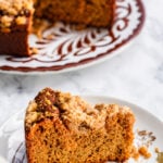 A slice of crumb-topped Sourdough Pumpkin Coffee Cake sits on a floral plate, with the rest of the cake on a larger patterned plate in the background. The cake appears moist with a golden-brown crumb.