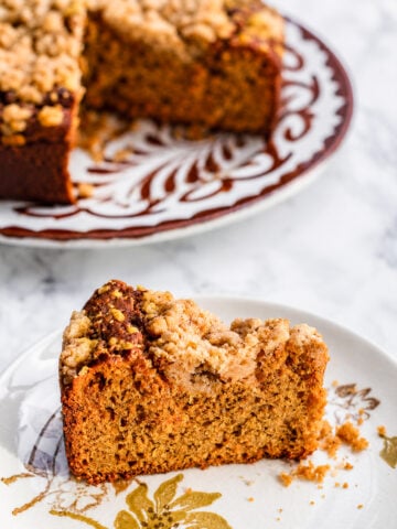 A slice of crumb-topped Sourdough Pumpkin Coffee Cake sits on a floral plate, with the rest of the cake on a larger patterned plate in the background. The cake appears moist with a golden-brown crumb.