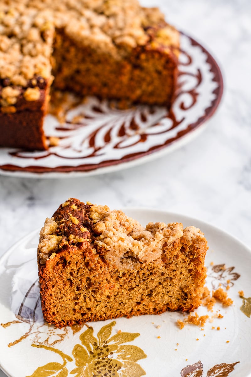 A slice of crumb-topped Sourdough Pumpkin Coffee Cake sits on a floral plate, with the rest of the cake on a larger patterned plate in the background. The cake appears moist with a golden-brown crumb.