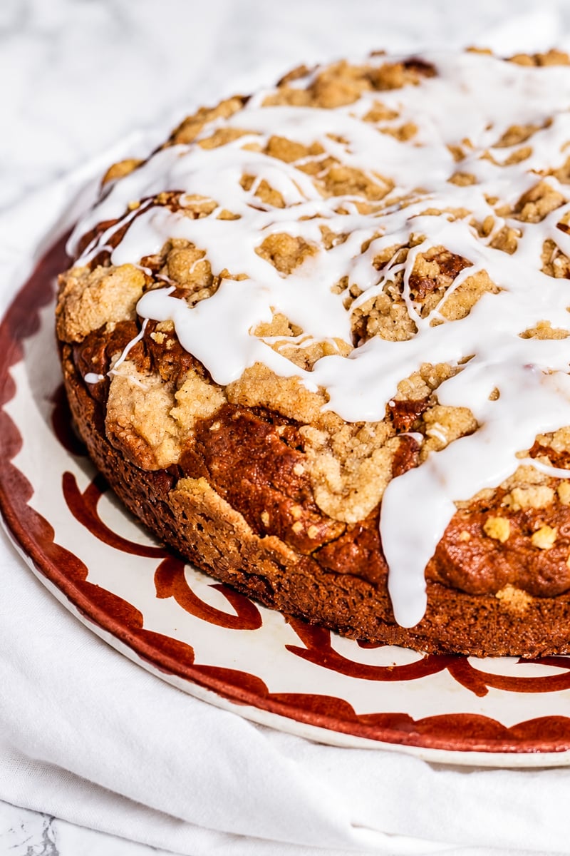 A round Sourdough Pumpkin Coffee Cake topped with white icing drizzle and crumbly streusel sits on a decorative plate, placed over a white cloth.