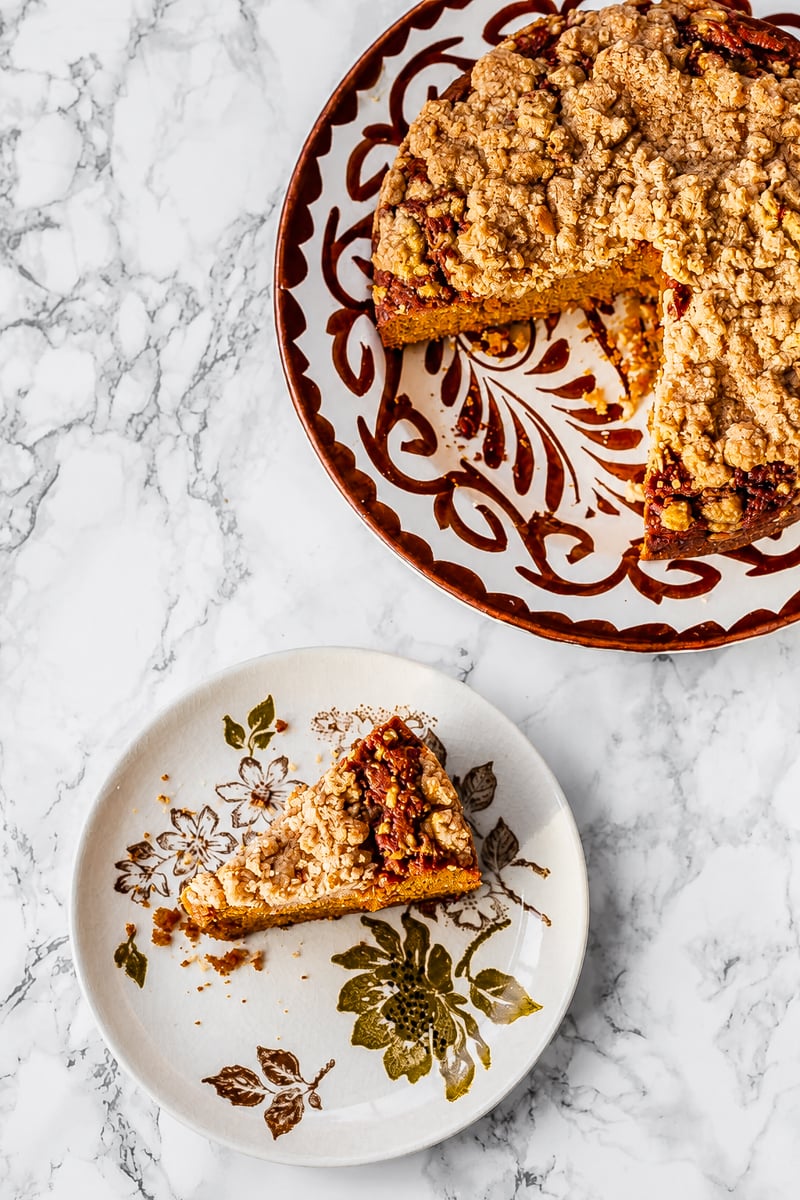 A round Sourdough Pumpkin Coffee Cake with a crumb topping sits on a decorative plate; a slice rests on a smaller floral plate nearby, all arranged on a marble surface.