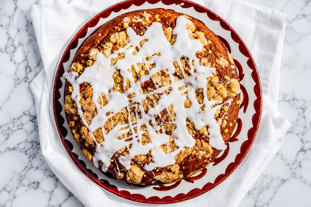 A round Sourdough Pumpkin Coffee Cake topped with crumbly streusel and white icing drizzle sits on a red-rimmed plate atop a white cloth and marble surface.