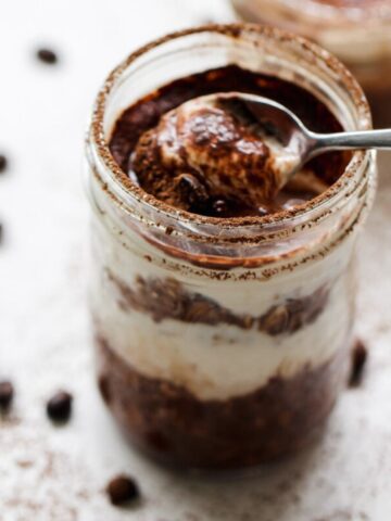 A spoon scoops creamy tiramisu oats from a glass jar, revealing distinct chocolate and cream layers. Coffee beans and cocoa powder are scattered on the white surface nearby.