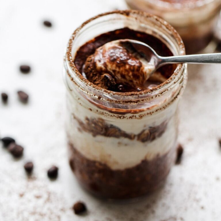A spoon scoops creamy tiramisu oats from a glass jar, revealing distinct chocolate and cream layers. Coffee beans and cocoa powder are scattered on the white surface nearby.