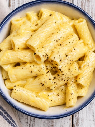 A bowl of creamy Vegan Cheese Sauce Pasta rigatoni topped with freshly ground black pepper, served in a blue-rimmed dish on a wooden table.