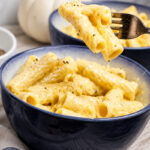 A close-up of creamy Vegan Cheese Sauce Pasta in a blue bowl, topped with black pepper. A fork lifts a bite, and a small white pumpkin is visible in the background.