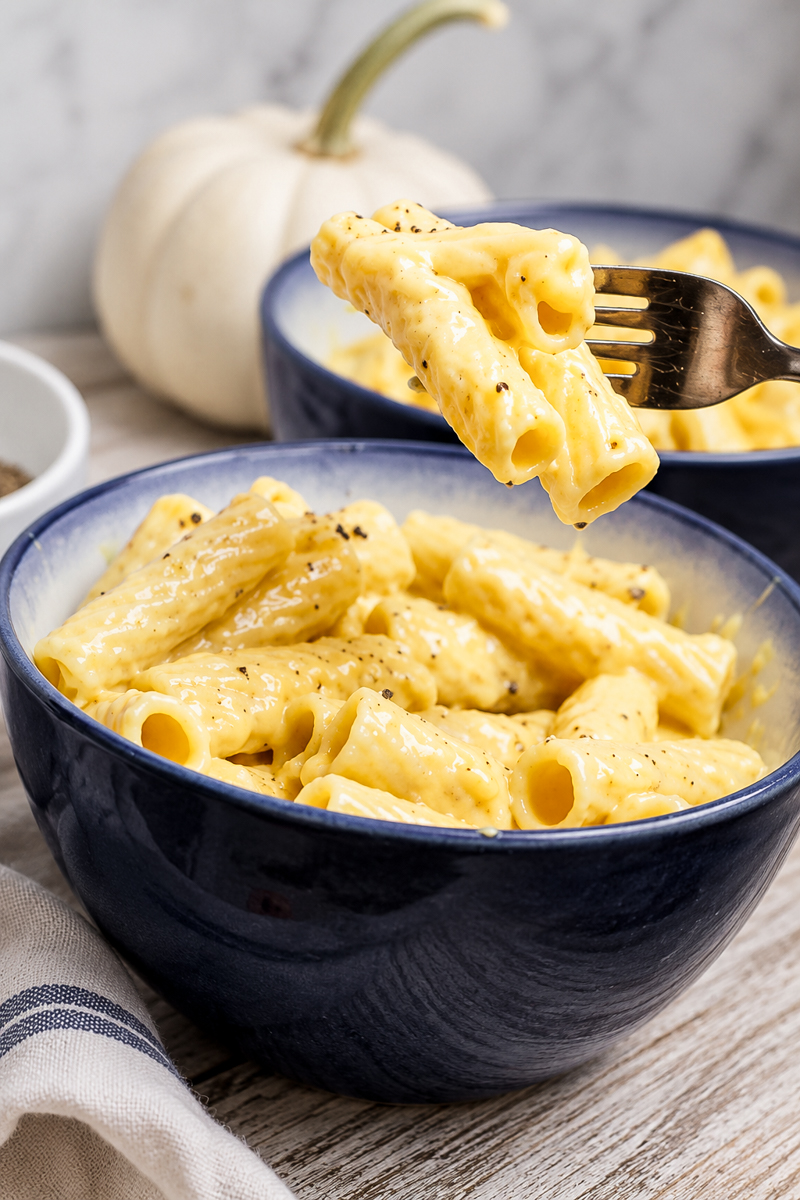 A close-up of creamy Vegan Cheese Sauce Pasta in a blue bowl, topped with black pepper. A fork lifts a bite, and a small white pumpkin is visible in the background.