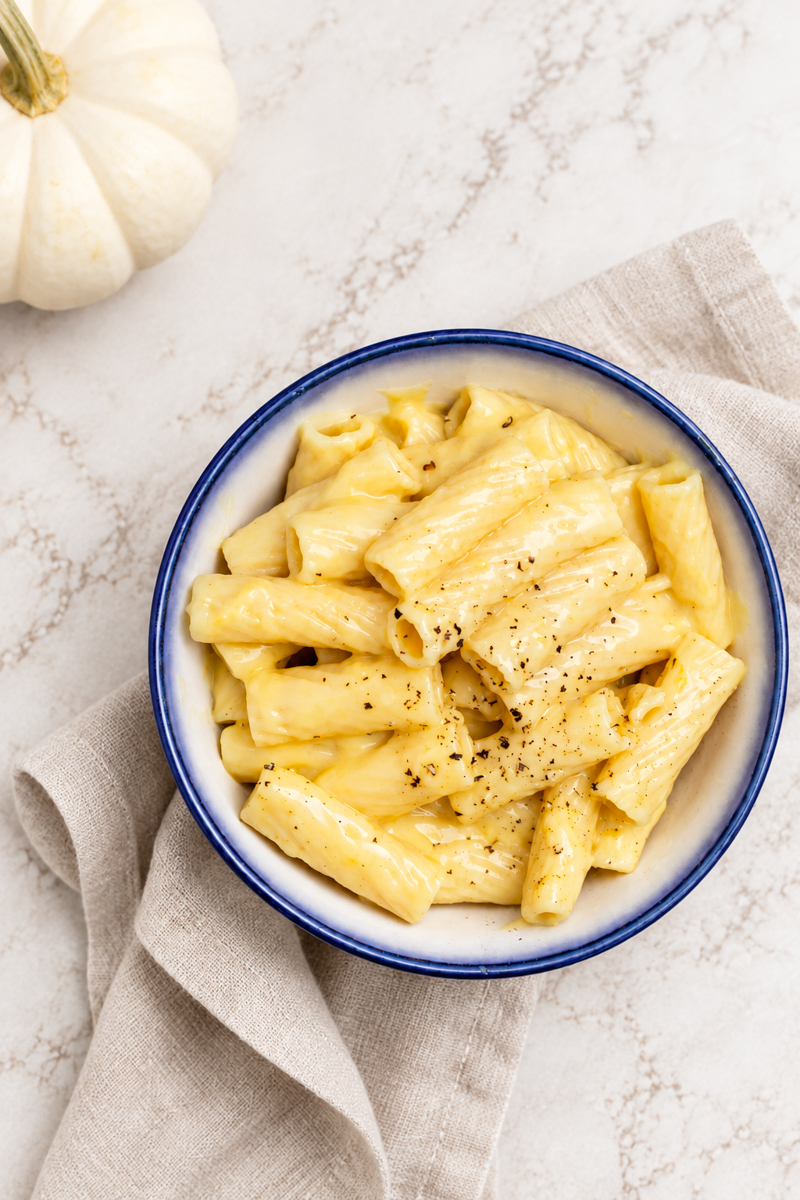 A bowl of creamy Vegan Cheese Sauce Pasta rigatoni topped with cracked black pepper sits on a beige napkin, with a white pumpkin visible in the upper left corner on a marble surface.