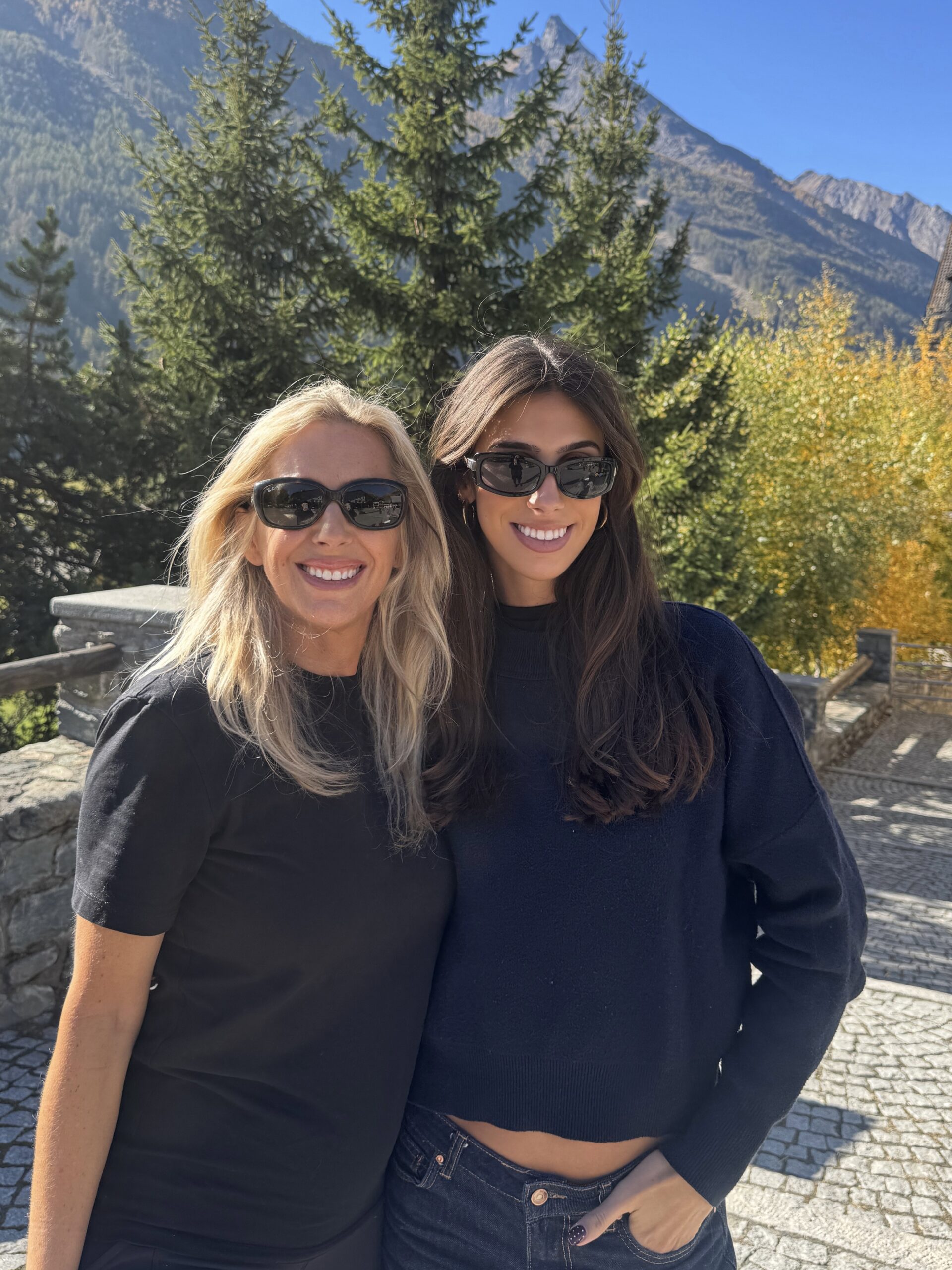 Two women wearing sunglasses and dark clothing stand smiling outdoors in front of trees and mountains on a sunny day. The sky is clear and blue, and autumn-colored foliage is visible in the background.