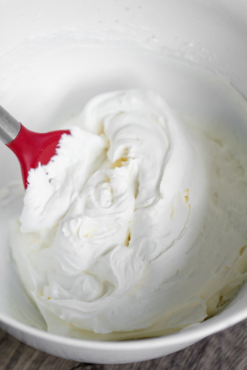 A close-up of fluffy whipped cream in a white mixing bowl, being stirred with a red and white spatula.