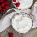 Two glass bowls of whipped cream on a wooden surface, one topped with three raspberries. A bowl of fresh raspberries and a white cloth are also nearby, with a few loose raspberries on the table.