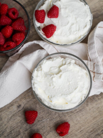 Two glass bowls of whipped cream on a wooden surface, one topped with three raspberries. A bowl of fresh raspberries and a white cloth are also nearby, with a few loose raspberries on the table.