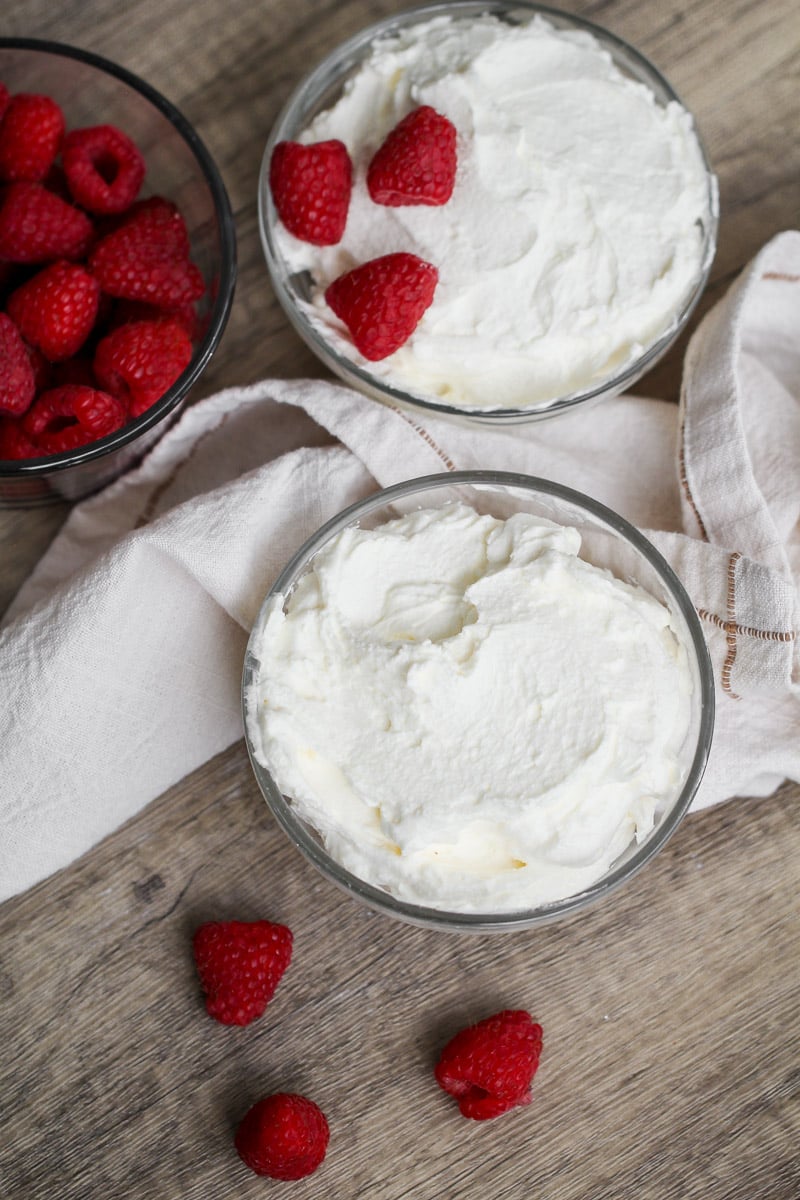 over head image of two glass bowls of whipped cream on a wooden surface, one topped with three raspberries. A bowl of fresh raspberries and a white cloth are also nearby, with a few loose raspberries on the table.