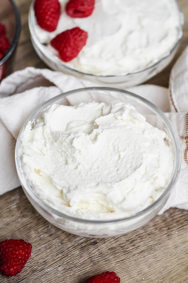 close up image of a glass bowl filled with whipped cream sits on a wooden surface, surrounded by a few fresh raspberries and a white cloth. Another bowl of whipped cream with raspberries is partially visible in the background.