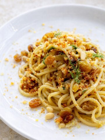 A plate of Pasta con le Sarde topped with golden raisins, pine nuts, breadcrumbs, herbs, and grated cheese, served on a white dish.