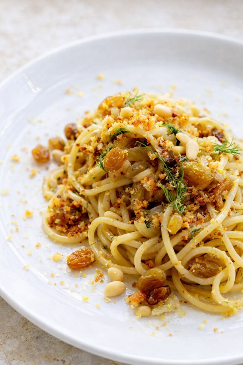 A plate of Pasta con le Sarde topped with golden raisins, pine nuts, breadcrumbs, herbs, and grated cheese, served on a white dish.