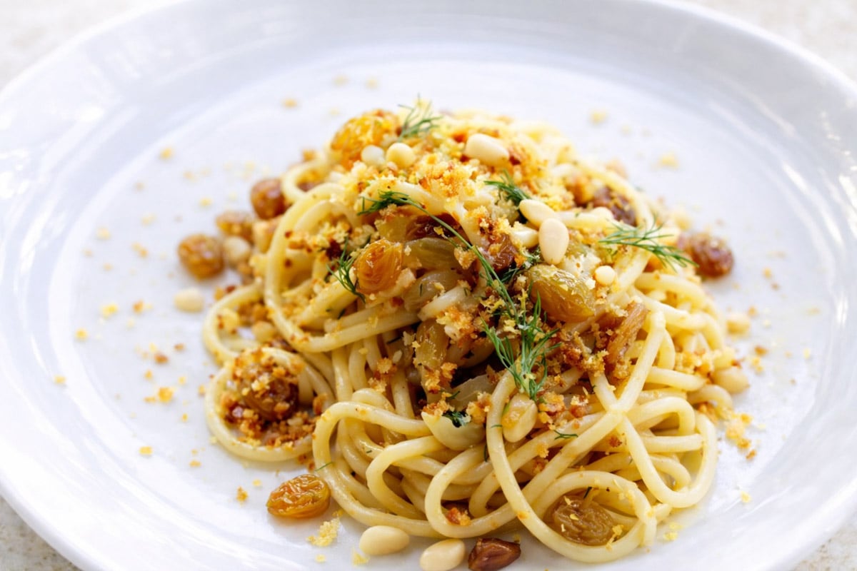 A plate of Pasta con le Sarde topped with golden raisins, pine nuts, fresh herbs, and breadcrumbs, presented on a white dish.