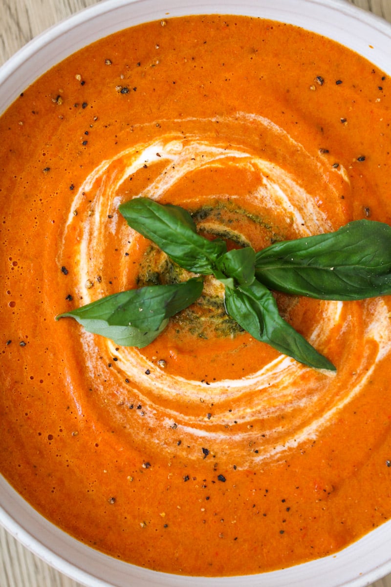 A bowl of creamy tomato soup with a swirl of cream, topped with fresh basil leaves and cracked black pepper, served on a light wooden surface.