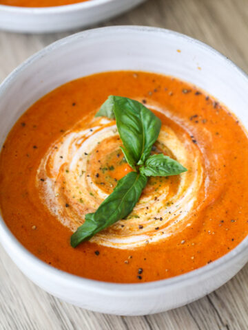 A bowl of creamy tomato soup garnished with a swirl of cream, freshly cracked black pepper, and a sprig of fresh basil, placed on a light wooden surface.