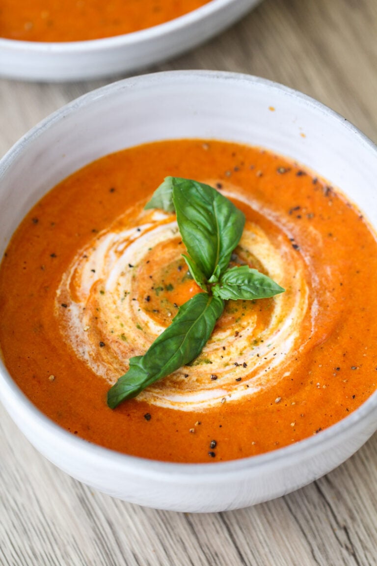 A bowl of creamy tomato soup garnished with a swirl of cream, freshly cracked black pepper, and a sprig of fresh basil, placed on a light wooden surface.
