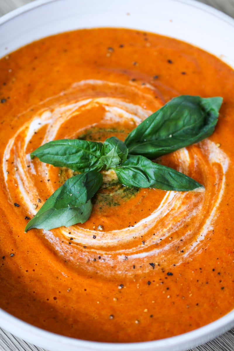 A close-up of creamy tomato soup with a swirl of cream and a garnish of fresh basil leaves in the center, served in a white bowl. Black pepper is sprinkled on top.