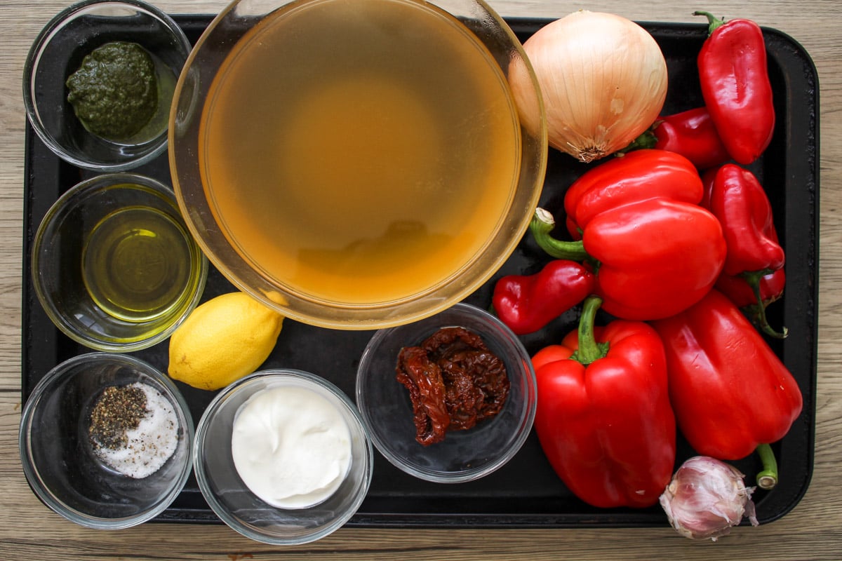 A tray with a bowl of broth, whole red bell peppers, an onion, a lemon, peppers, a garlic bulb, sun-dried tomatoes, creme fraiche, pesto, olive oil, and a small bowl of salt and pepper.