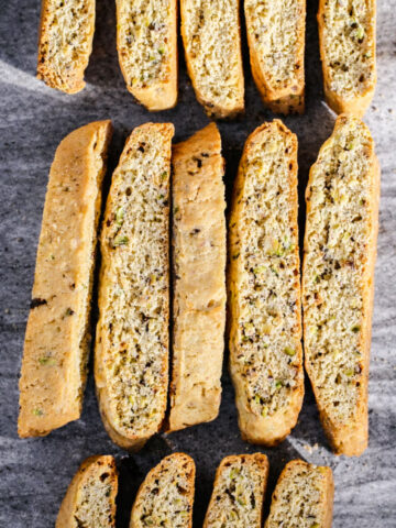 Rows of golden-brown biscotti with visible pieces of nuts are arranged neatly on a gray stone surface, showing their crisp texture and speckled interior.