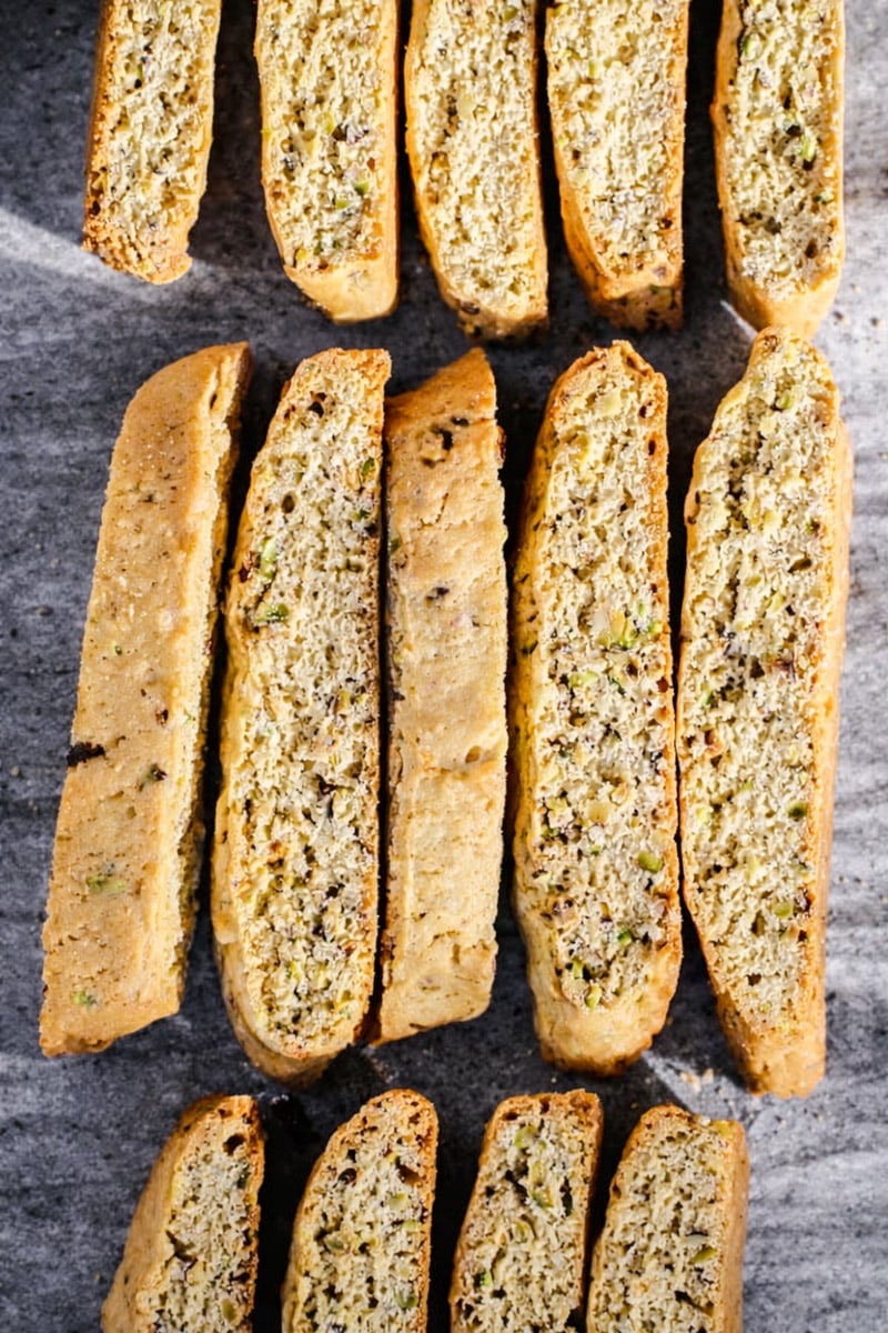 Rows of golden-brown biscotti with visible pieces of nuts are arranged neatly on a gray stone surface, showing their crisp texture and speckled interior.