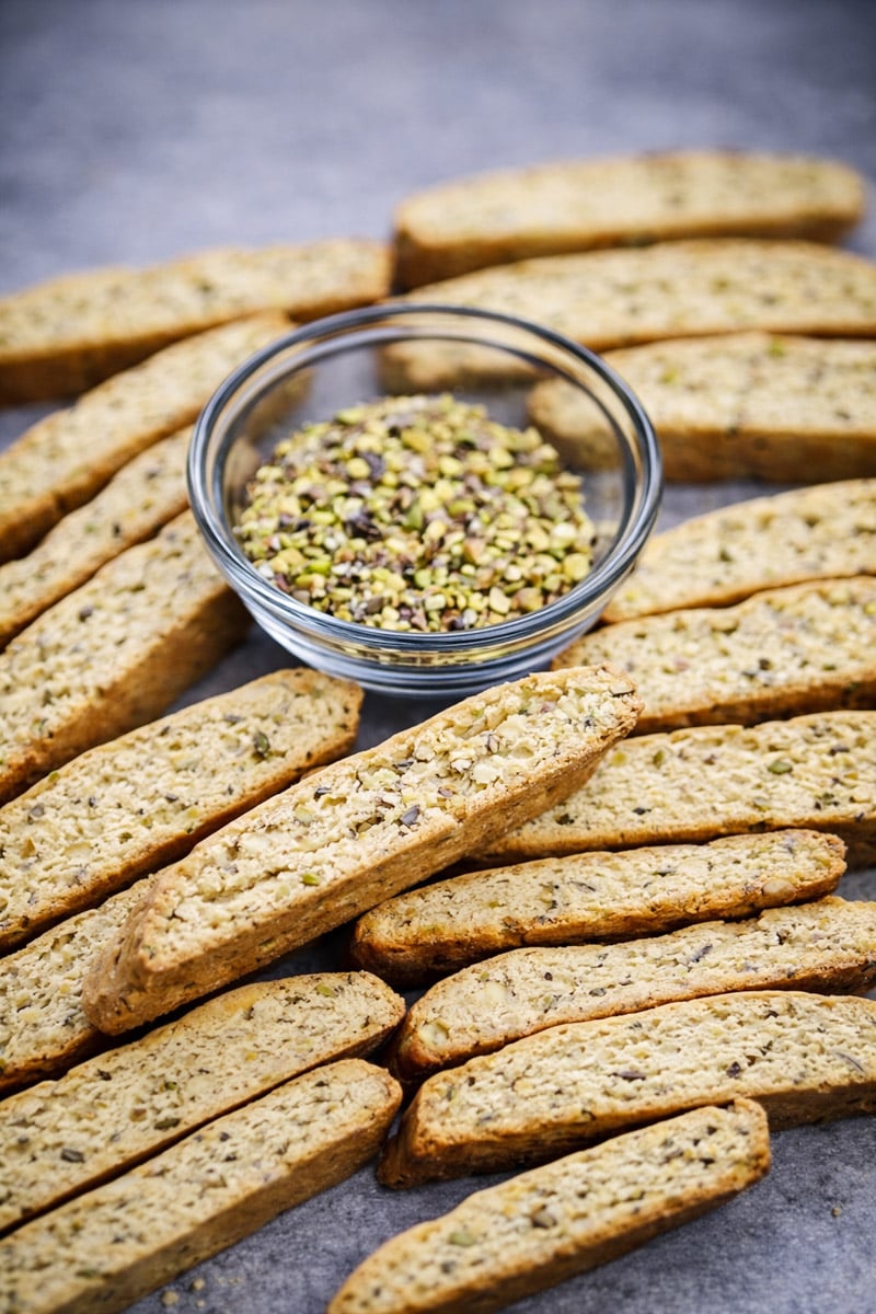 A bowl of chopped pistachios is surrounded by several pieces of pistachio biscotti on a gray surface.