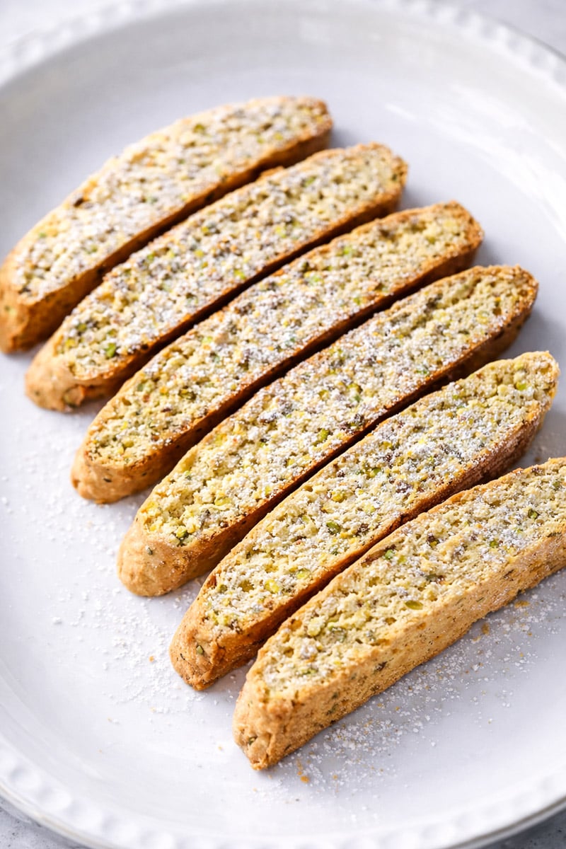 Six pieces of biscotti are arranged in a row on a white plate, lightly dusted with powdered sugar. The biscotti appear golden brown and have a textured surface.