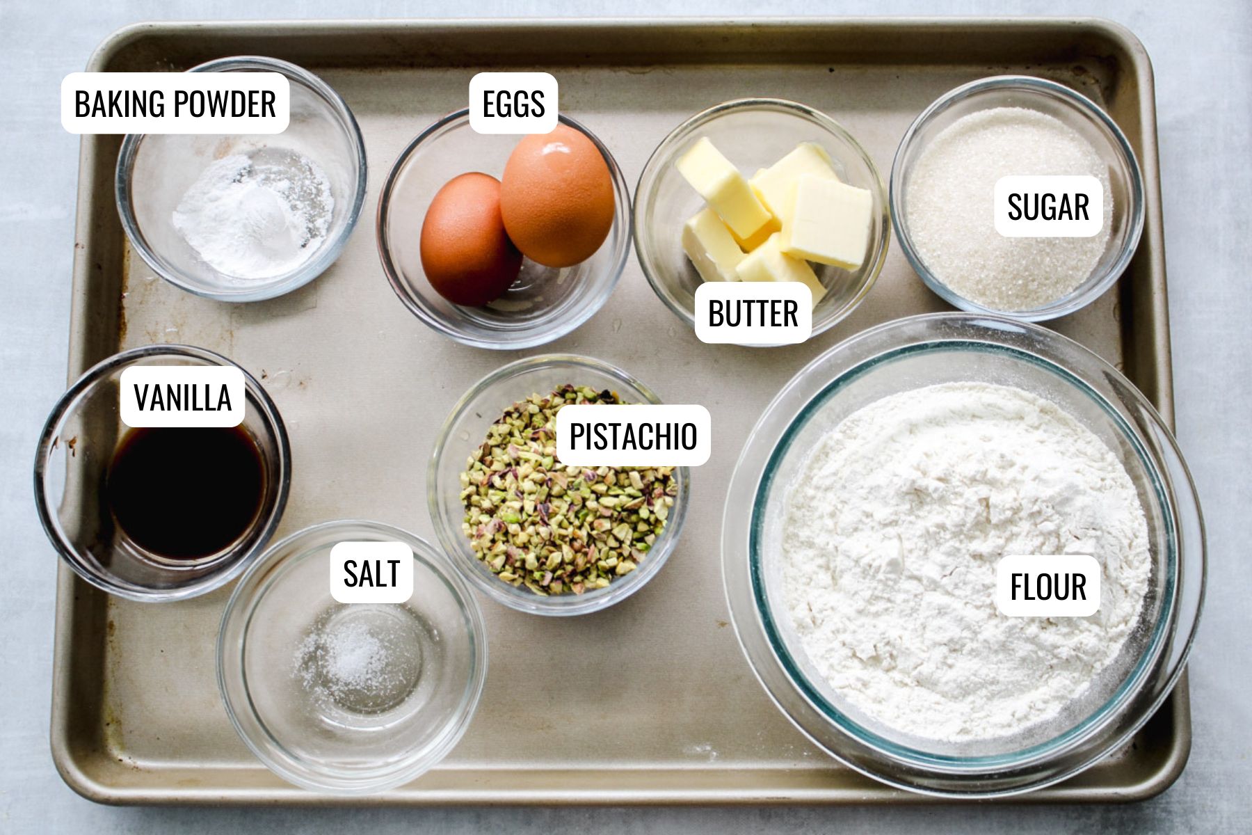 A baking tray holds small bowls labeled and filled with baking powder, eggs, butter, sugar, vanilla, salt, chopped pistachios, and flour, all organized neatly for baking preparation.