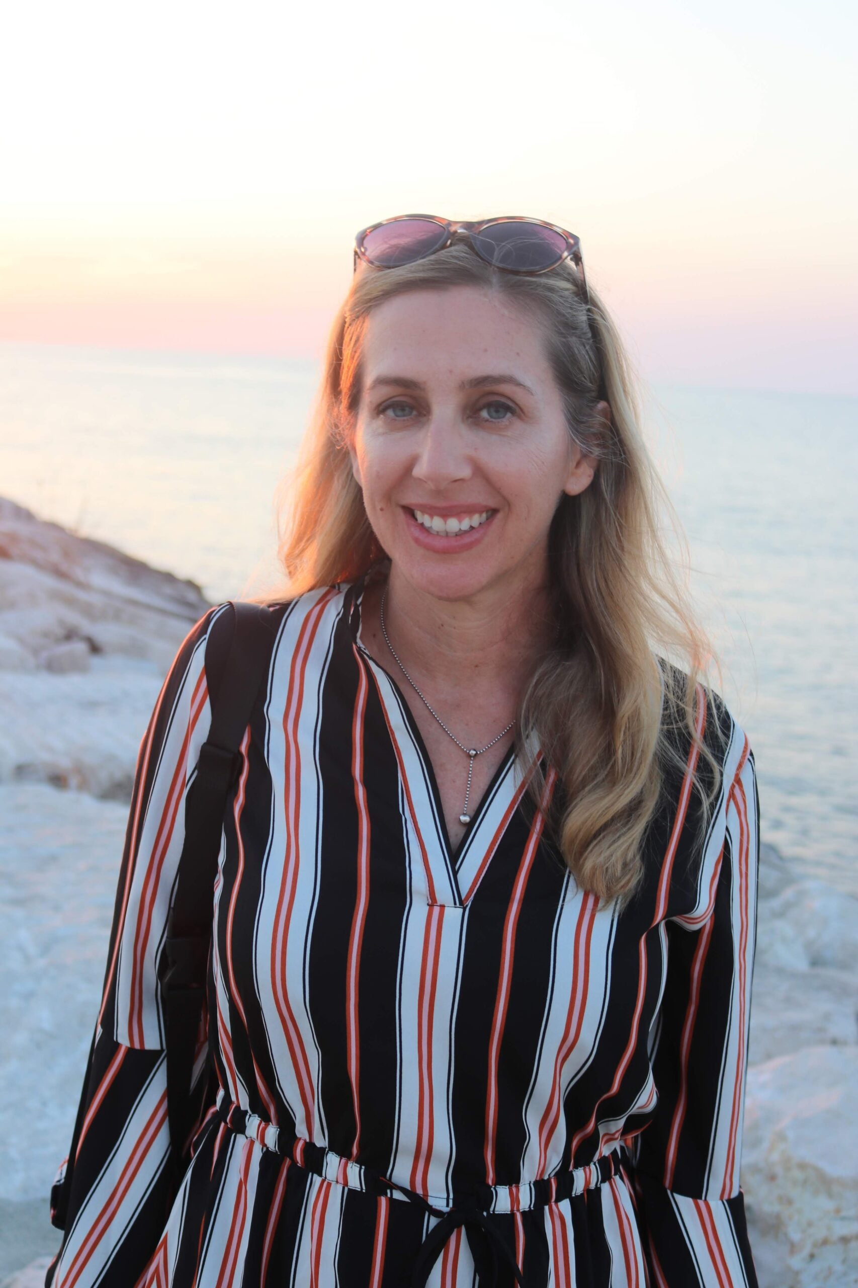 A woman with long blonde hair, wearing sunglasses on her head and a striped dress, smiles at the camera while standing on rocky terrain near the sea at sunset during her Italy trip planning.