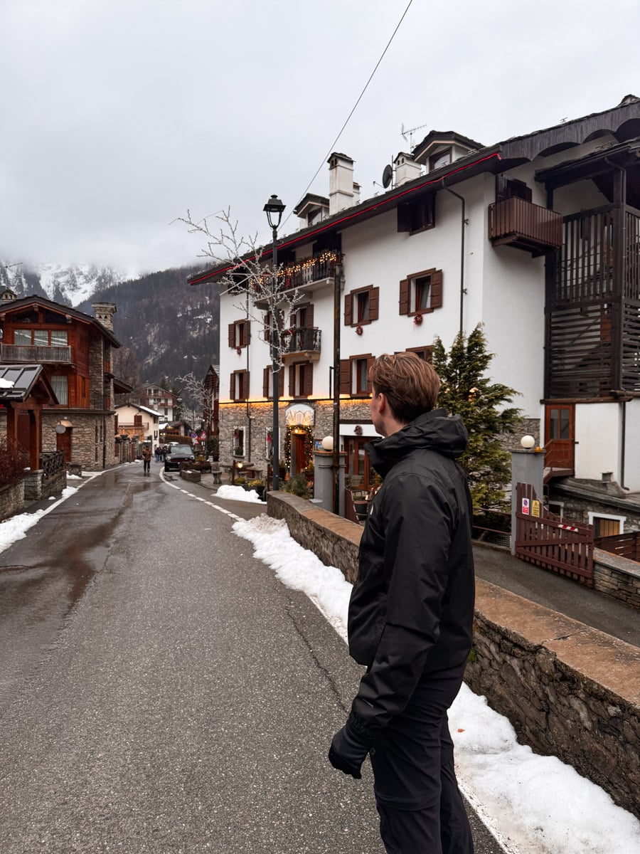 A man in a black jacket stands on a snowy street in a quaint mountain village with alpine-style buildings, brown shutters, and snow-capped mountains visible under a cloudy sky.