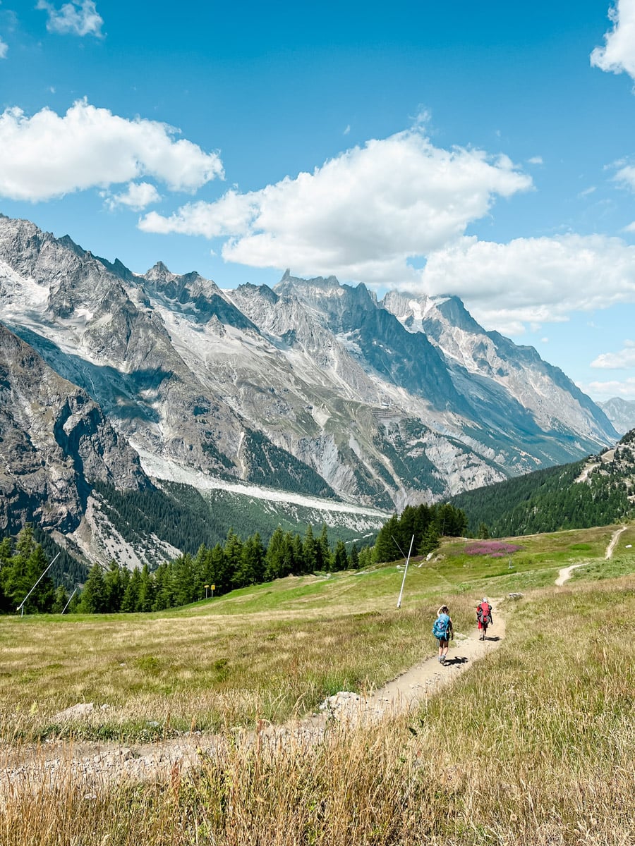 Three hikers walk along a dirt trail through a grassy alpine meadow with tall, rocky mountains and patches of snow in the background under a blue sky with scattered clouds.
