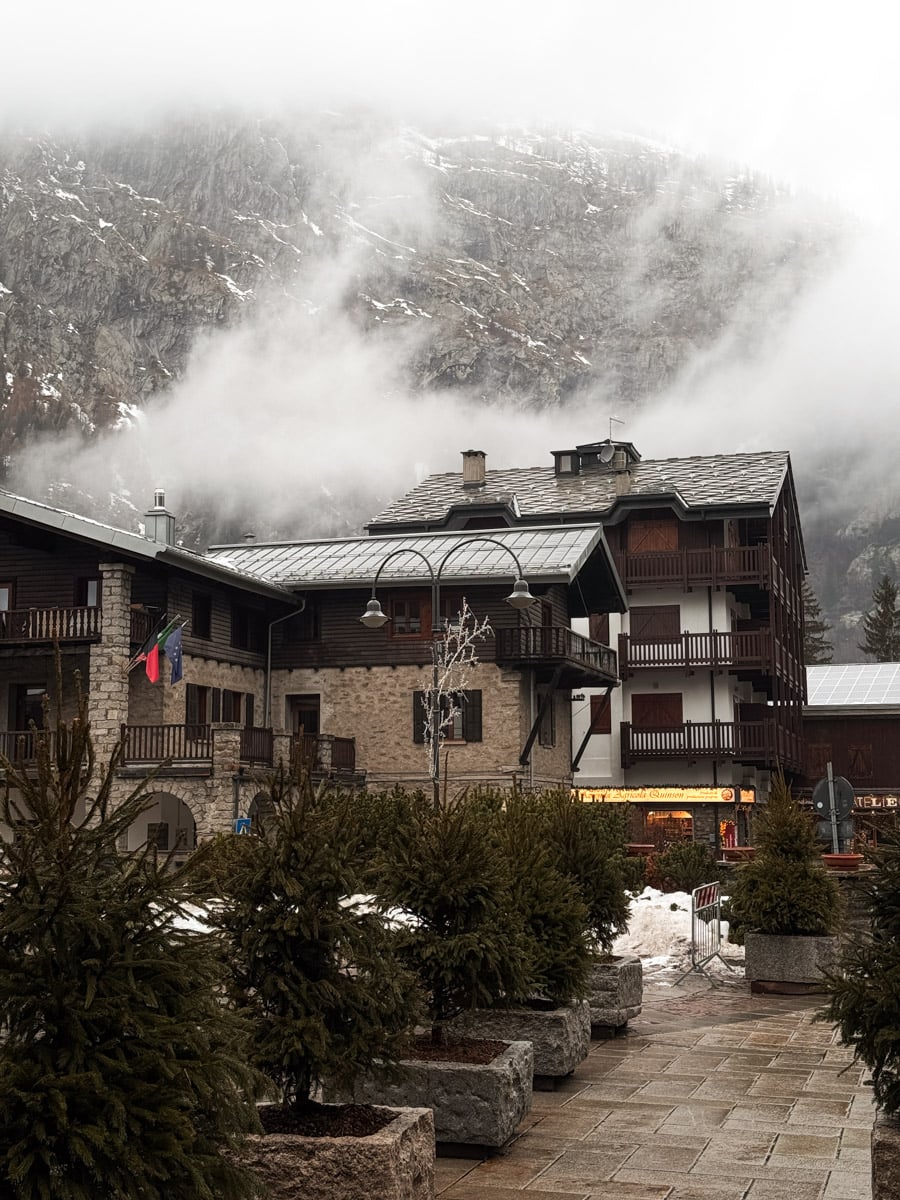 Mountain village scene with stone and wood buildings, fir trees, and a cloudy, misty mountain in the background. Snow patches are visible on the ground, and the atmosphere appears cold and wintry.