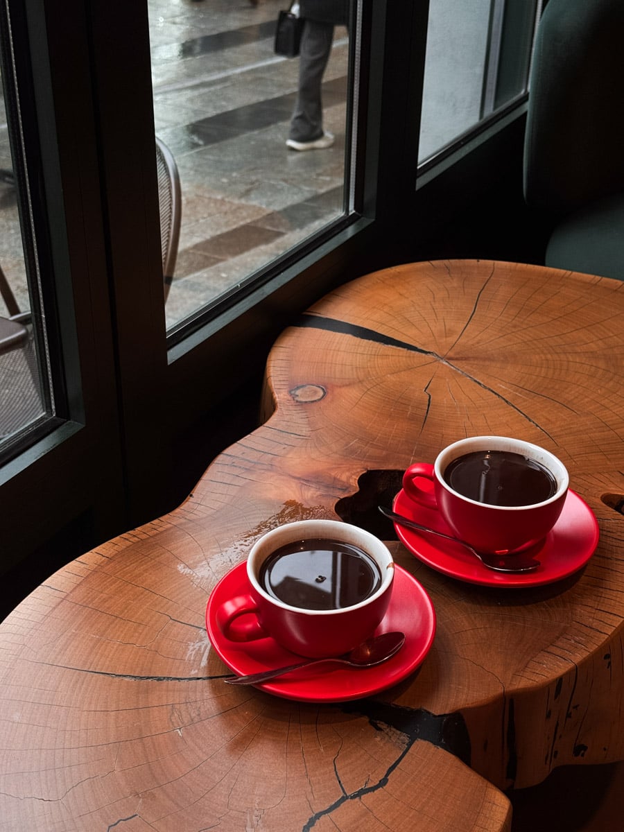 Two red cups of coffee sit on a rustic wooden table by a window. Outside, a person walks past on a rainy day, visible through the glass. The atmosphere appears cozy and inviting.