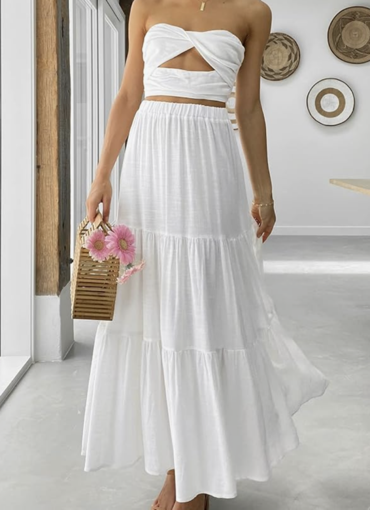 A woman wearing a white, strapless, two-piece maxi dress-an ideal choice for what to wear in Italy-holds a wooden handbag with pink flowers. She stands barefoot indoors near large windows and decorative wall plates.