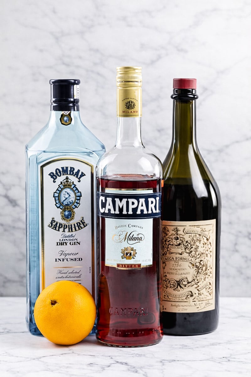 Three bottles&mdash;Bombay Sapphire gin, Campari, and a bottle of vermouth&mdash;stand side by side on a white marble surface, with a whole orange placed in front of them.
